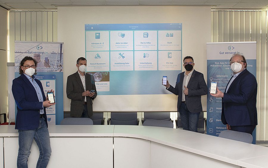 Das Bild zeigt 4 Männer mit Mund-Nasen-Bedeckungen, die Smartphones in den Händen halten. Im Hintergrund wird der Startbildschirm der neuen App auf eine Leinwand übertragen.