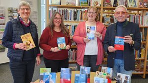 4 Personen halten Bücher hoch, davor ein Tisch mit weiteren Büchern. Hinter ihnen sind Bücherregale zu sehen