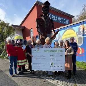 8 Frauen, 1 Mann und eine Person im Maus-Kostüm und Feuerwehrmantel posieren mit übergroßen Spendenscheck