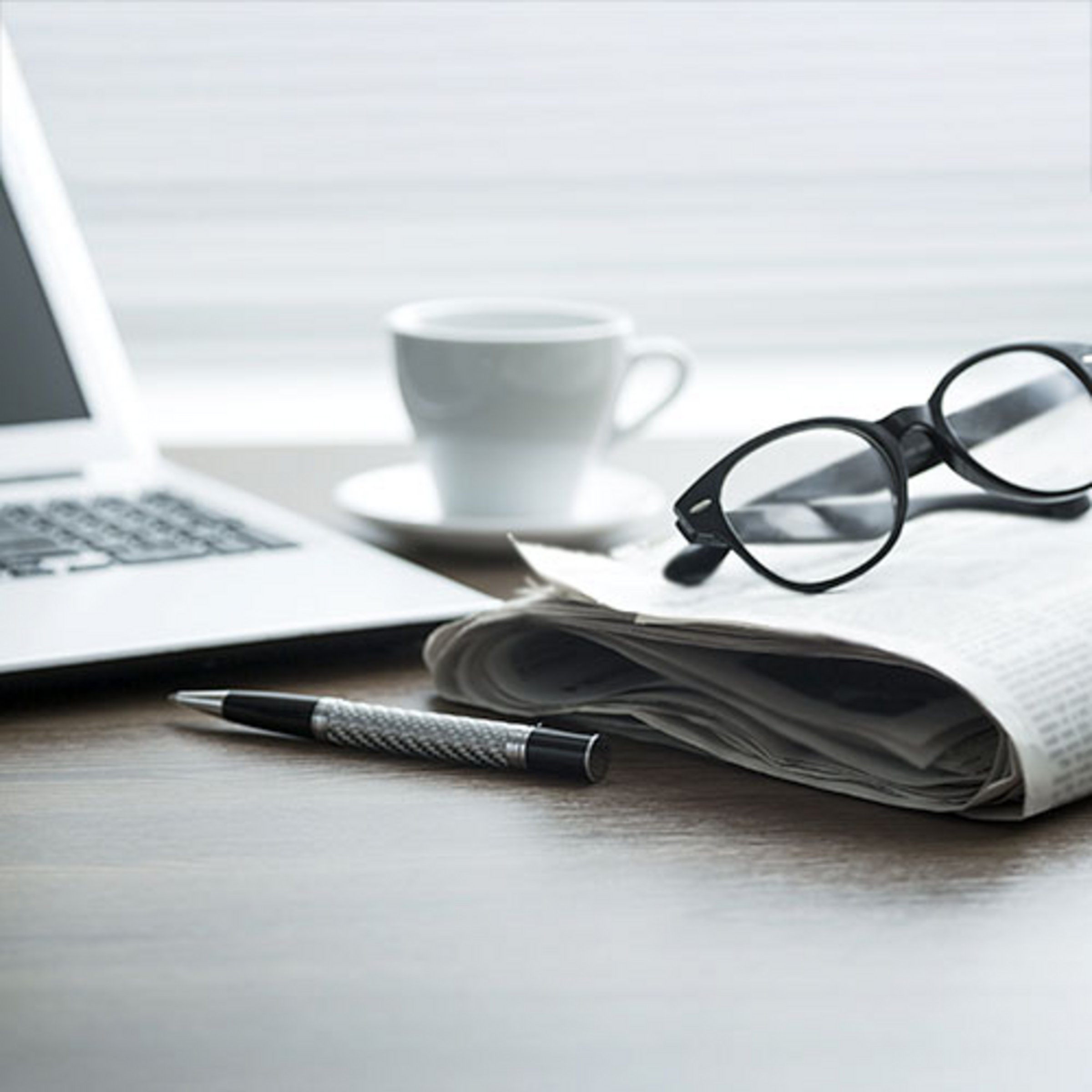 Das Bild zeigt einen Laptop, eine Zeitung, eine Kaffeetasse, eine Brille und einen Stift.
