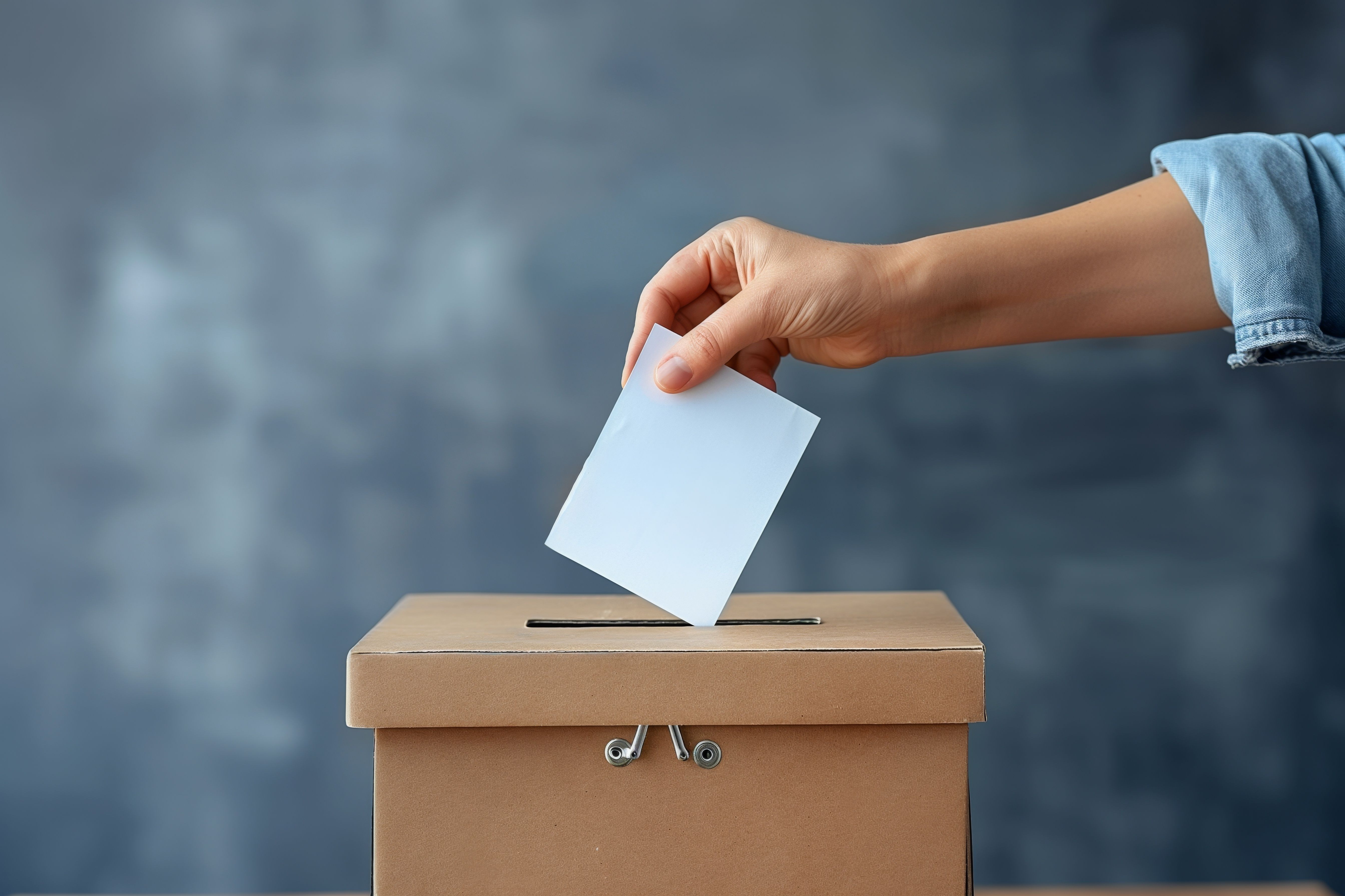 Das Bild zeigt eine Person, die einen Wahlzettel in eine Wahlurne wirft vor blau-grauem Hintergrund.